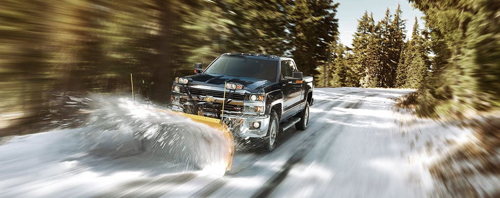 black chevrolet silverado with plow attachment on as it drives down a snowy road