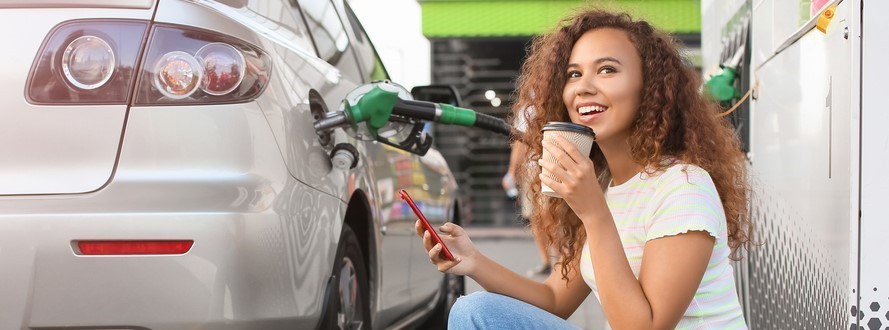 young woman sits on curb, smiling with coffee and cell phone in hand, next to gas pump while filling up her car