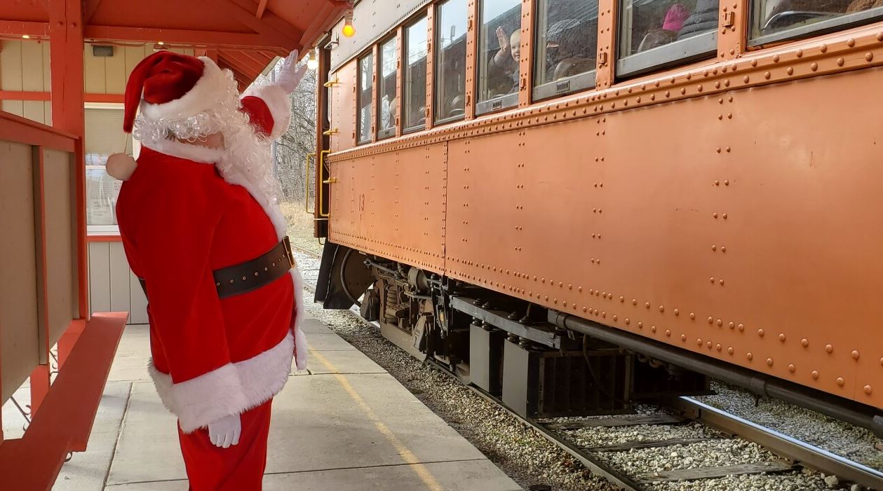at a train stop, Santa waives at children on a red train car