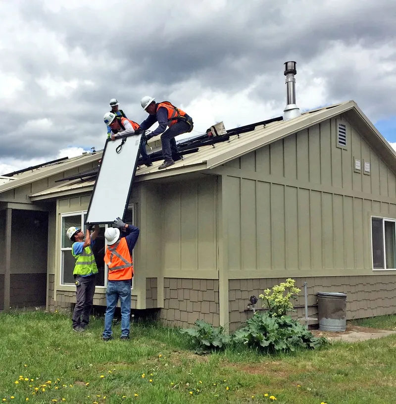 Workers in hard hats and safety vests lifting a solar panel onto a residential roof.