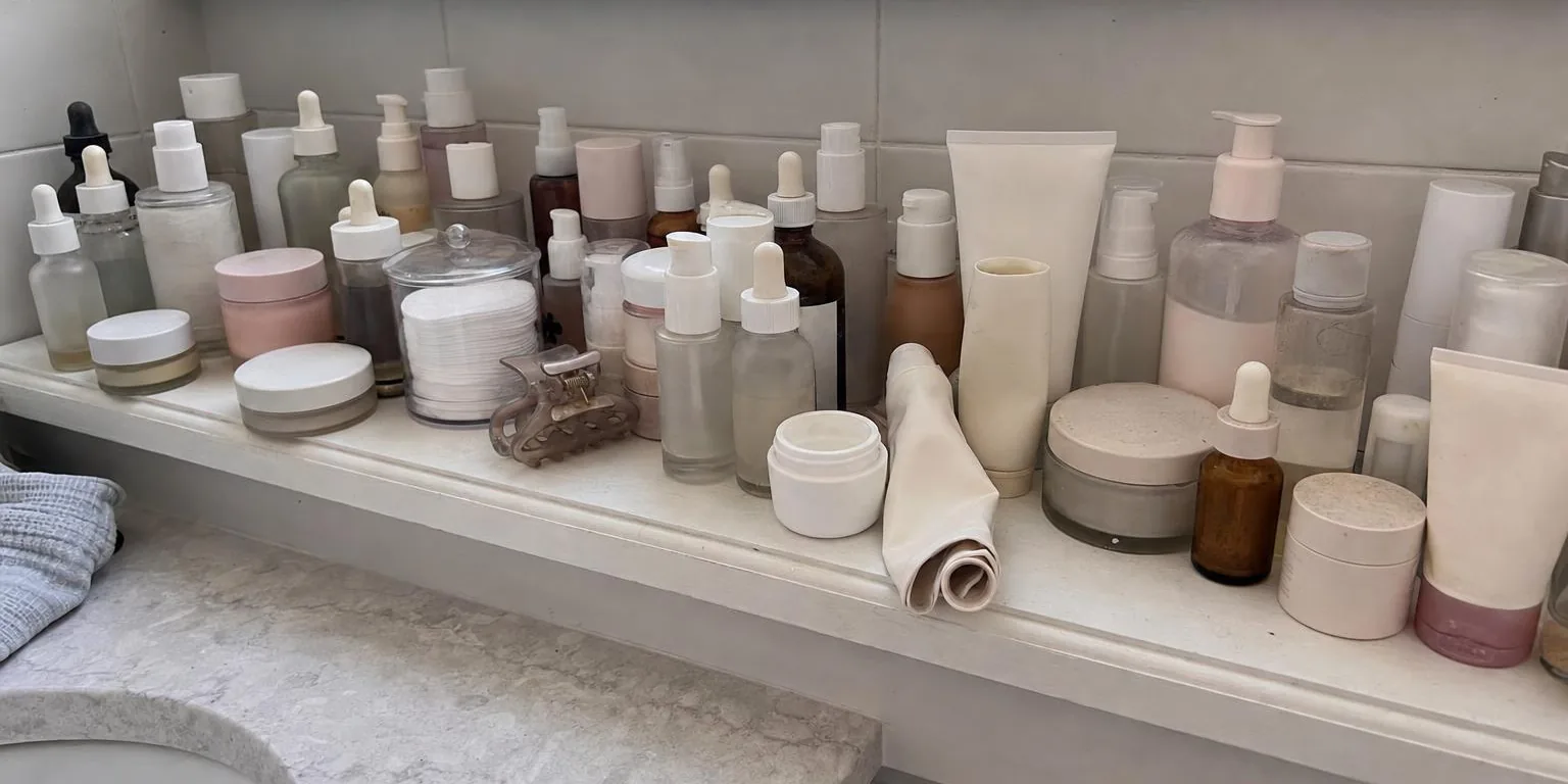 A crowded bathroom shelf packed with half-used skincare bottles and jars, some with dust on the lids