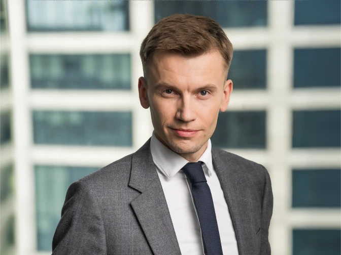 A man with short brown hair and a confident expression, wearing a grey textured suit jacket, white shirt, and navy blue tie. He is posed in front of a blurred background of high-rise office windows.