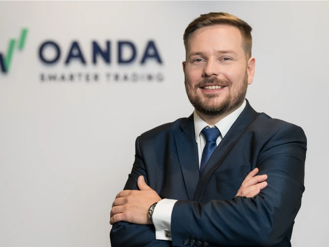 A man with short brown hair and a beard, smiling with his arms crossed while wearing a navy blue suit and tie. He is posed in front of a white wall with a blurred OANDA 'Smarter Trading' logo.