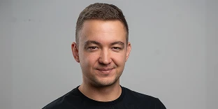 A man with short brown hair and a friendly, subtle smile, wearing a black crew-neck shirt against a plain grey background.