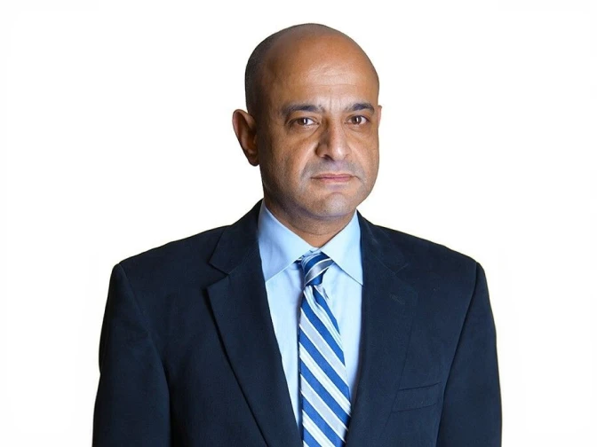 Portrait of a bald man with a serious, professional expression, wearing a dark suit jacket, a light blue shirt, and a blue and white striped tie. The background is a solid, neutral white.