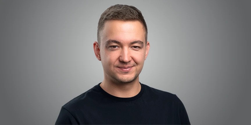 A man with short brown hair and a friendly, subtle smile, wearing a black crew-neck shirt against a plain grey background.