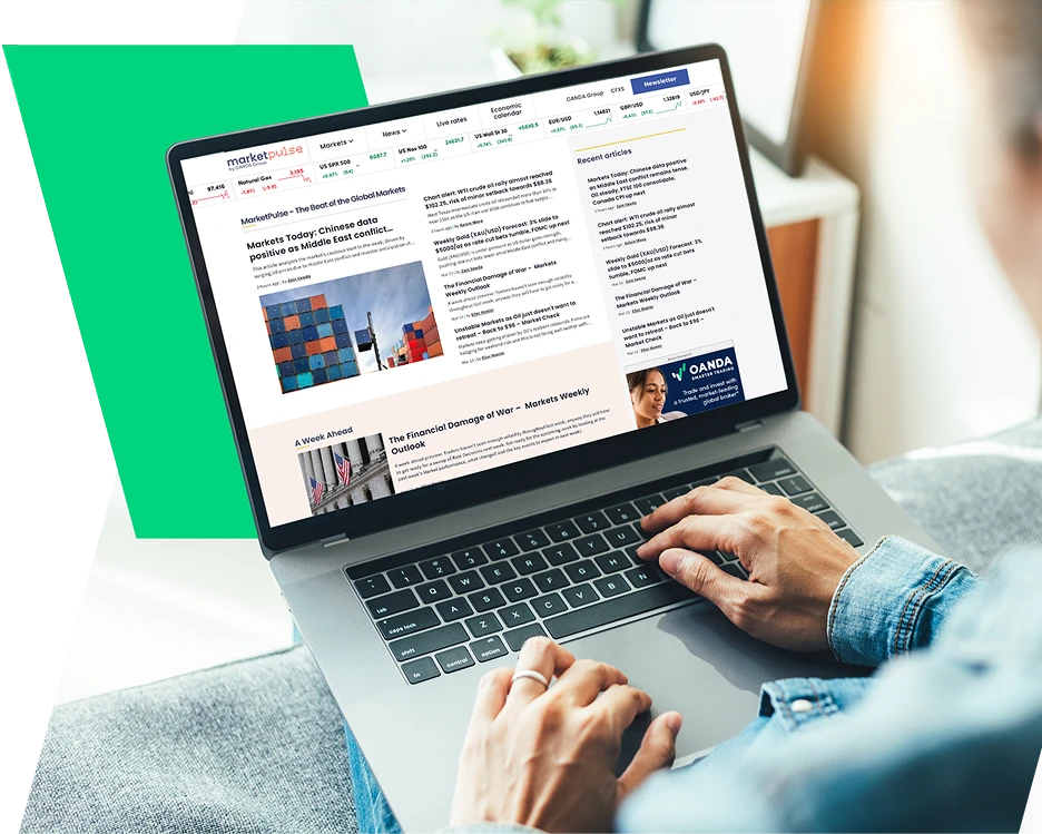 A person typing on an open laptop that is resting on their lap. The laptop screen displays a financial news website titled "MarketPulse by OANDA," featuring articles about global markets, oil prices, and economic outlooks. The person is wearing a denim sh
