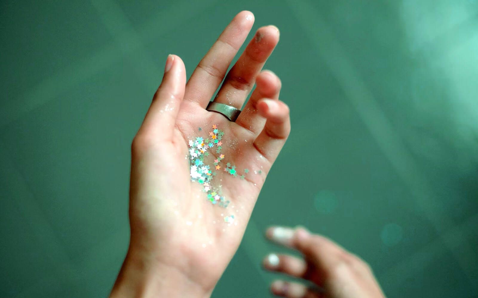 Women's hands with a ring on the middle finger holding very tiny shimmering stars of various colors.