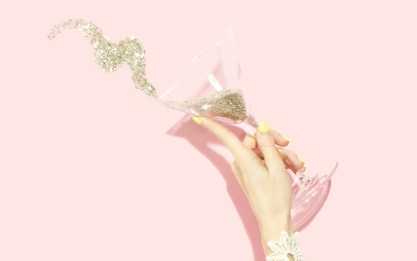 Women's hands with yellow nails, on a pink background, pouring the contents of a martini glass, inside the glass are finely cut shimmering golden stars.