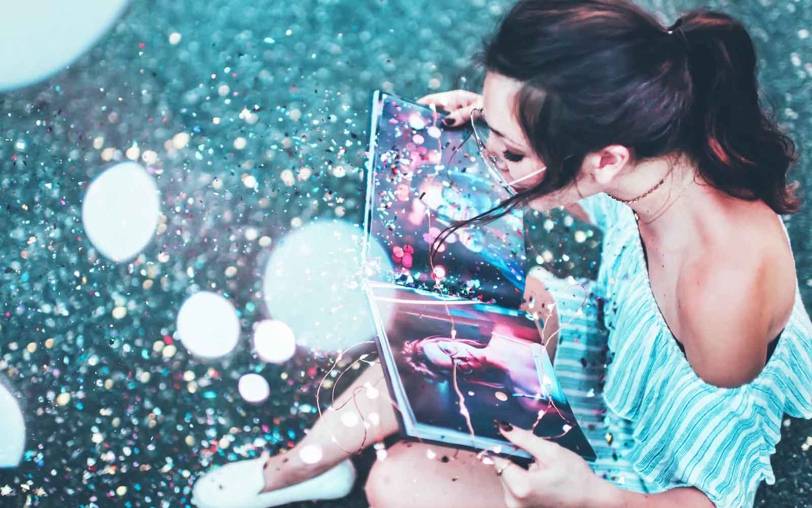 A girl with glasses opens a photobook with her picture and a strong gust of air blows away the glitter from the photobook.