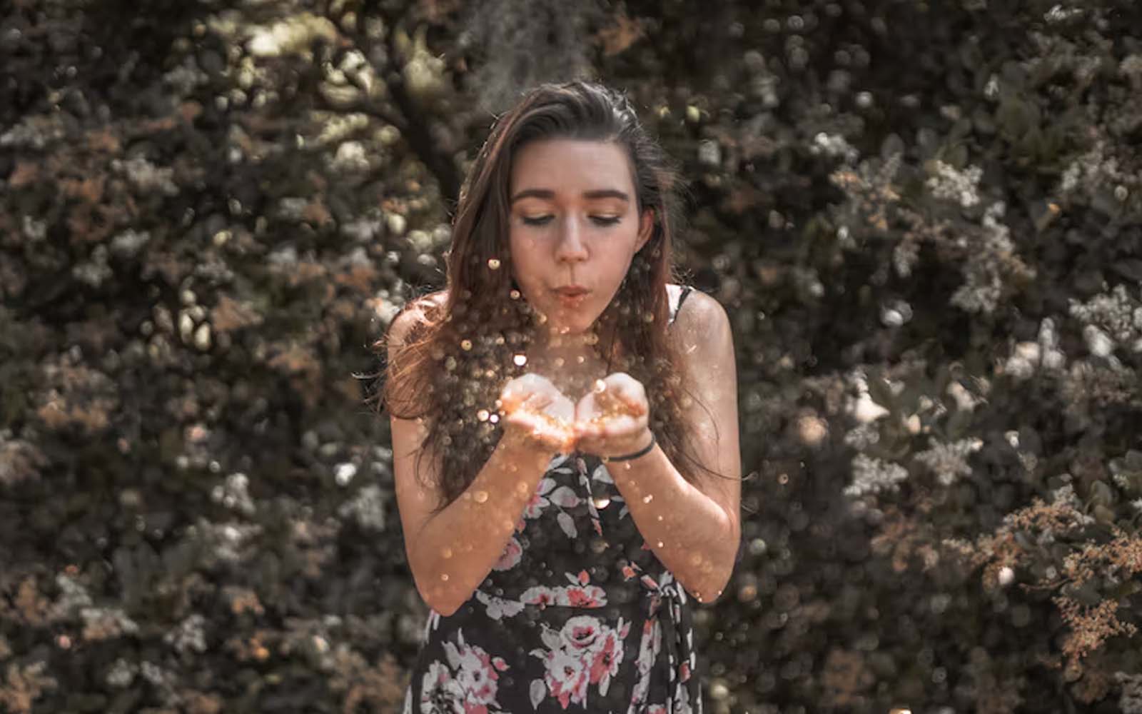 A girl with long hair against a natural backdrop blows into her palms, and glitter disperses around.