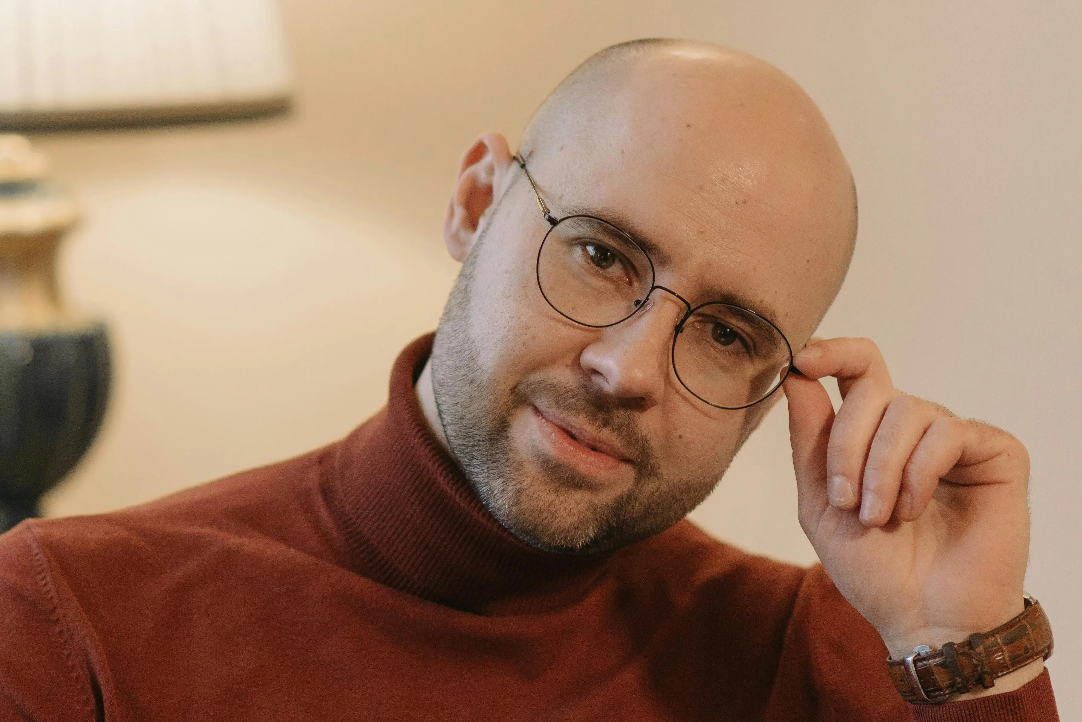 A man in a red sweater looking at the camera while holding the edge of his eyeglasses.