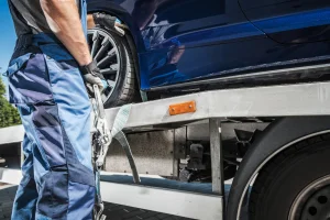 Tow truck driver loading a blue car with auto breakdown protection on the back of a flat bed tow truck