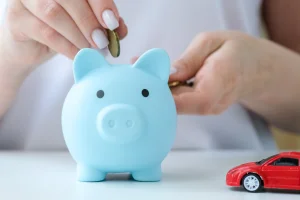 Hand putting coin in blue piggy bank next to toy red car saving money to buy a budget friendly warranty