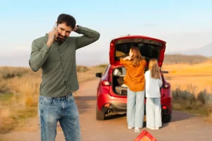 Man with powertrain warranty coverage on a cell phone in front of a broken down car red car with family on the way to vacation