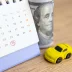 Photo illustration of a monthly car warranty payment with a calendar, roll of $100 bills, and a toy car