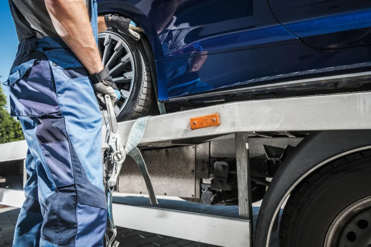Tow truck driver loading a blue car with auto breakdown protection on the back of a flat bed tow truck