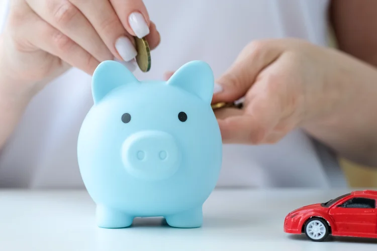Hand putting coin in blue piggy bank next to toy red car saving money to buy a budget friendly warranty