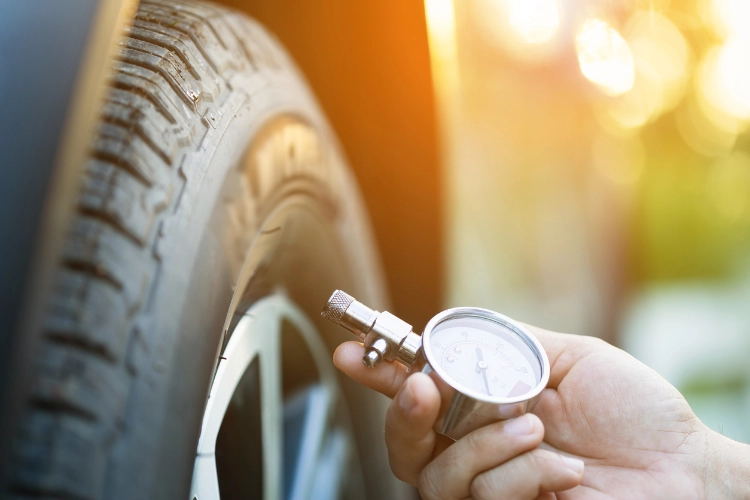 Close-up of a hand checking tire pressure with a gauge, with sunlight highlighting the tread of the tire.