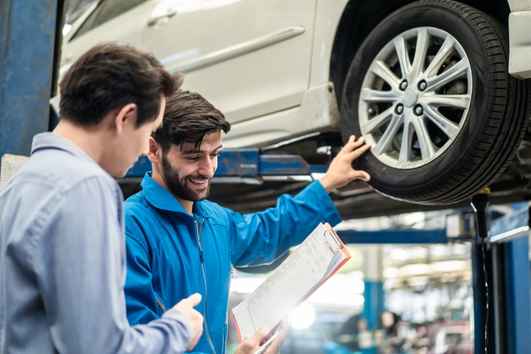 Mechanic showing a clipboard with a service checklist to a customer while inspecting a vehicle raised on a lift in an auto repair shop.