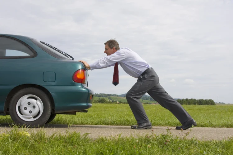 man in a tie pushing a green compact car that has broken down