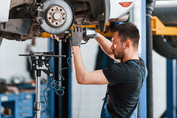 Auto mechanic using a power tool to repair a car's brake system while the vehicle is lifted on a hydraulic lift in a professional garage.