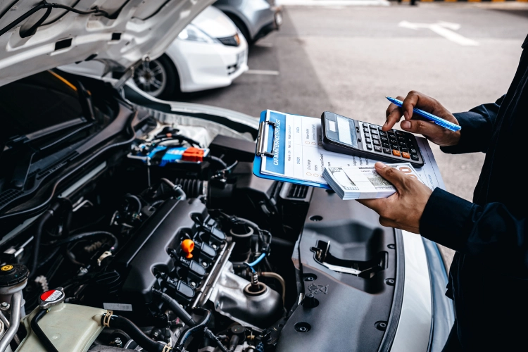 A mechanic calculating car repair costs with a clipboard and calculator next to an open vehicle hood in a service area.