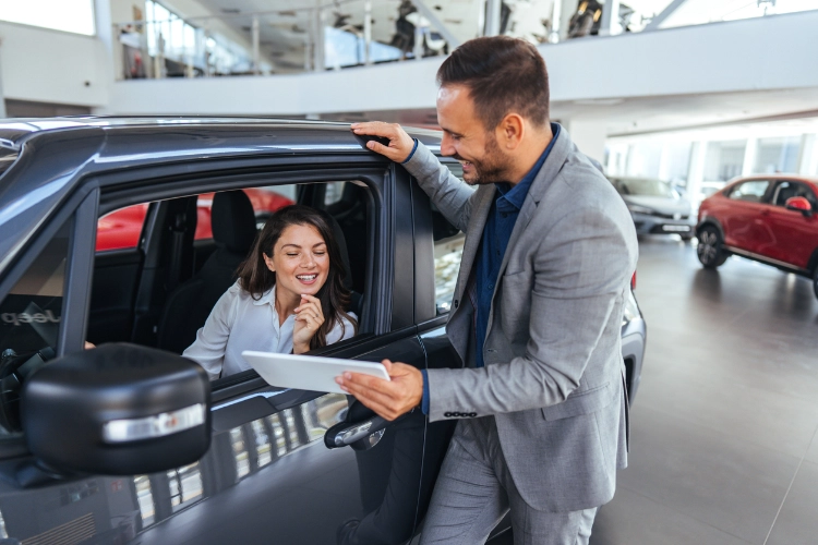 Smiling woman sitting in a rental car, speaking with an employee holding a tablet while her personal vehicle is being serviced.