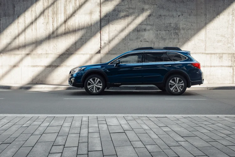 Side profile view of Subaru Outback on a sunny day, shadows and light beams on grey wall in the back.