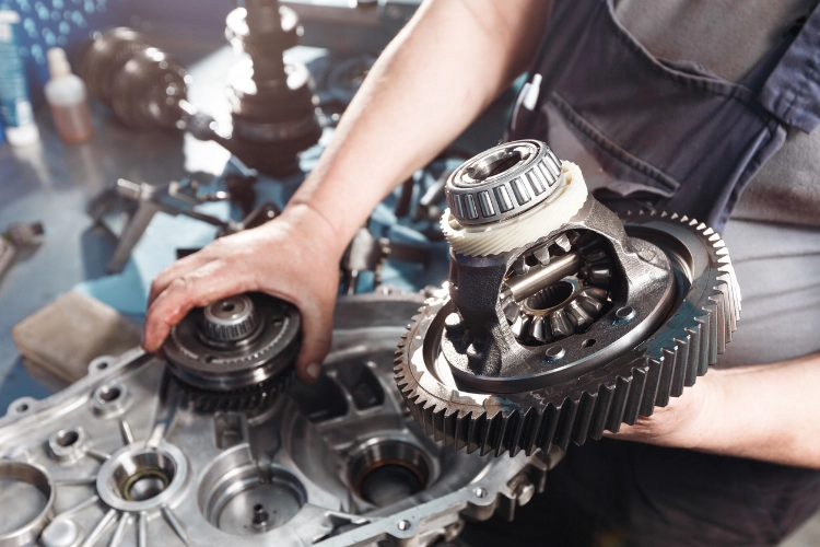 Close-up of a mechanic working on a disassembled transmission, holding a differential gear assembly at a workshop bench.