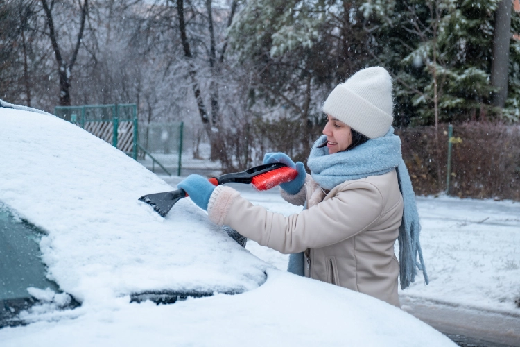 Woman bundled in winter clothing using a snow brush to clear ice from her car windshield on a snowy day.