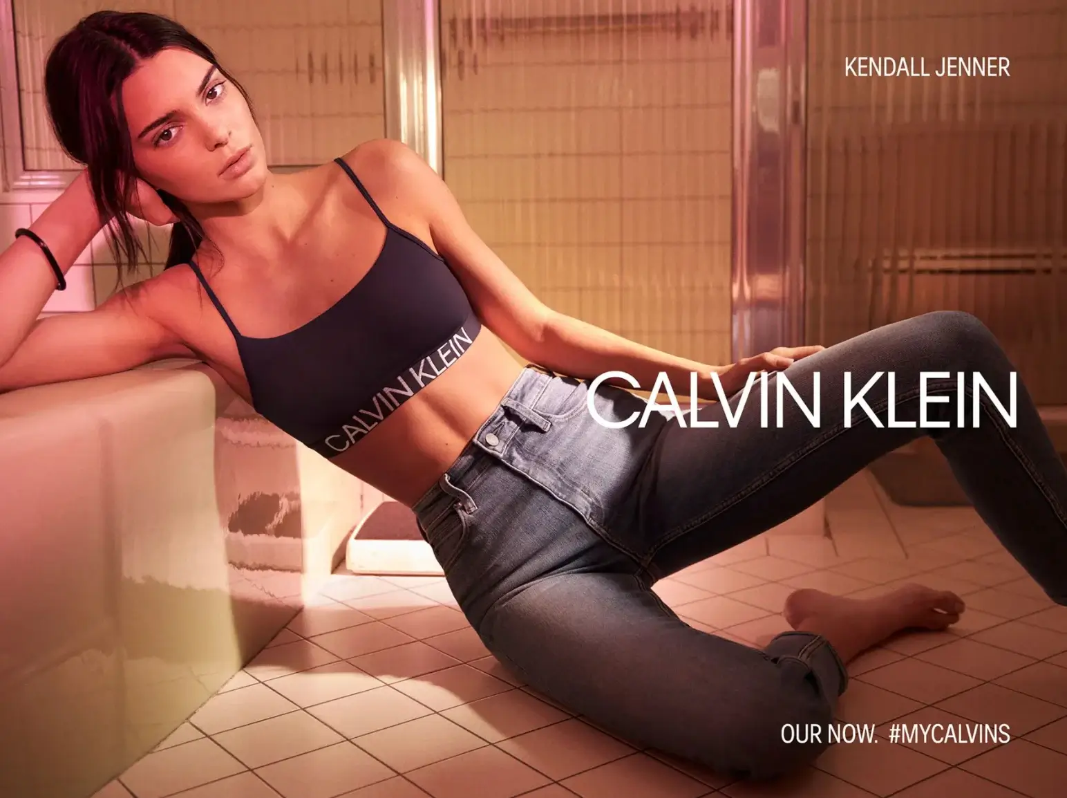 This image shows a woman, who leans against a tiled wall wearing a dark Calvin Klein sports bra and high-waisted jeans. Text on the image includes "CALVIN KLEIN" and the hashtag "#MYCALVINS" alongside "OUR NOW."