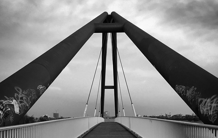 A high-contrast black and white photograph looking down the center of a pedestrian bridge. Two massive, converging structural beams frame the viewpoint, leading the eye with power and strength toward a city skyline on the distant horizon. The bridge floor provides a stable path, flanked by railings. This geometric composition of the bridge and its cables symbolizes Douglas USA's innovative, transformative services and its 