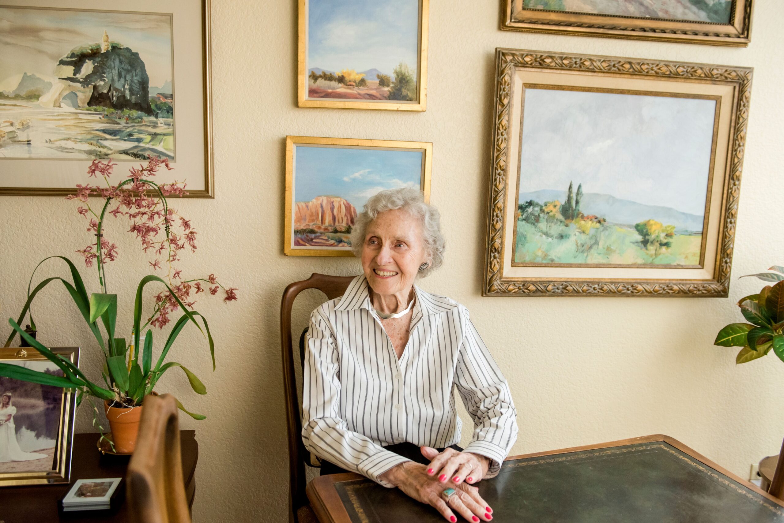 A senior woman sits smiling at a table with various watercolor paintings hanging on the wall behind her.