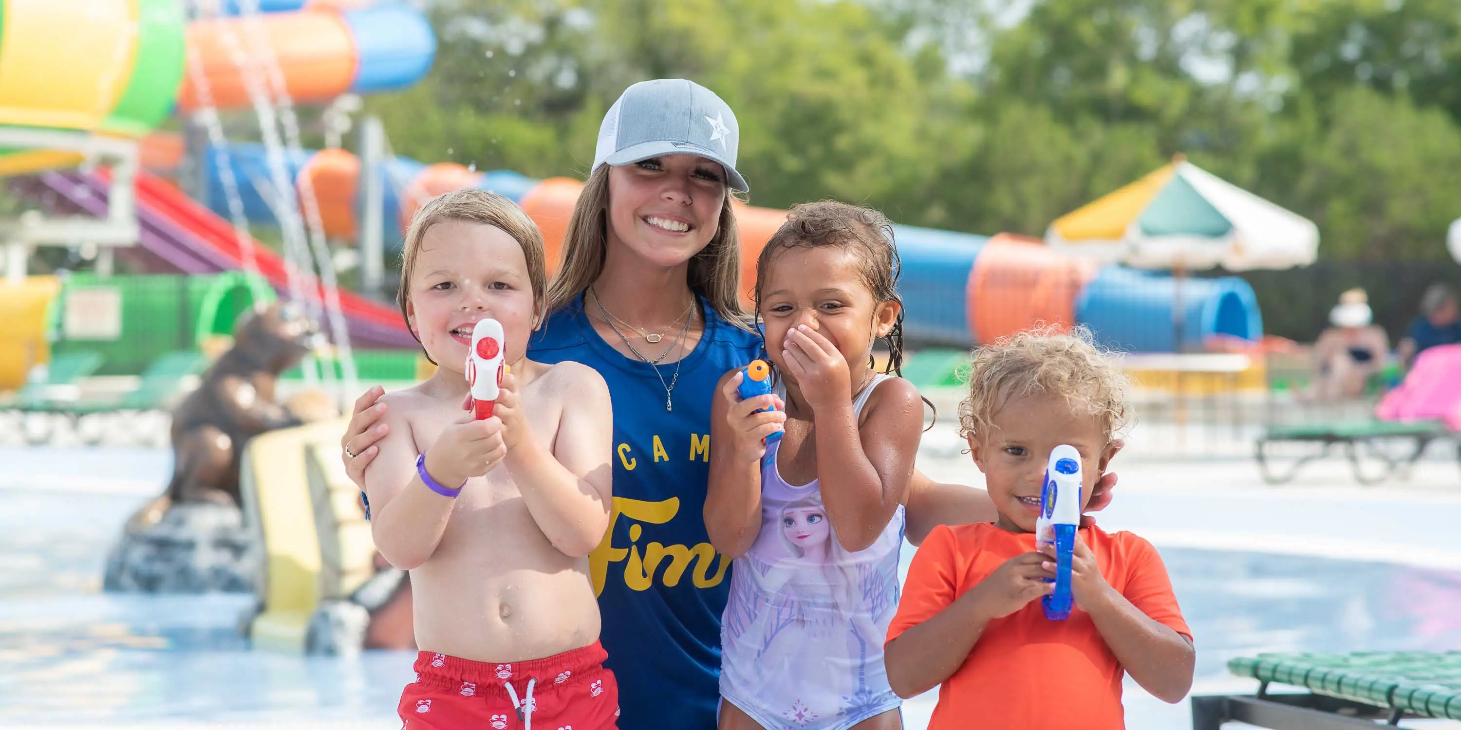 A Camp Fimfo staff member at the water playground, posing with children that are playing with squirt guns