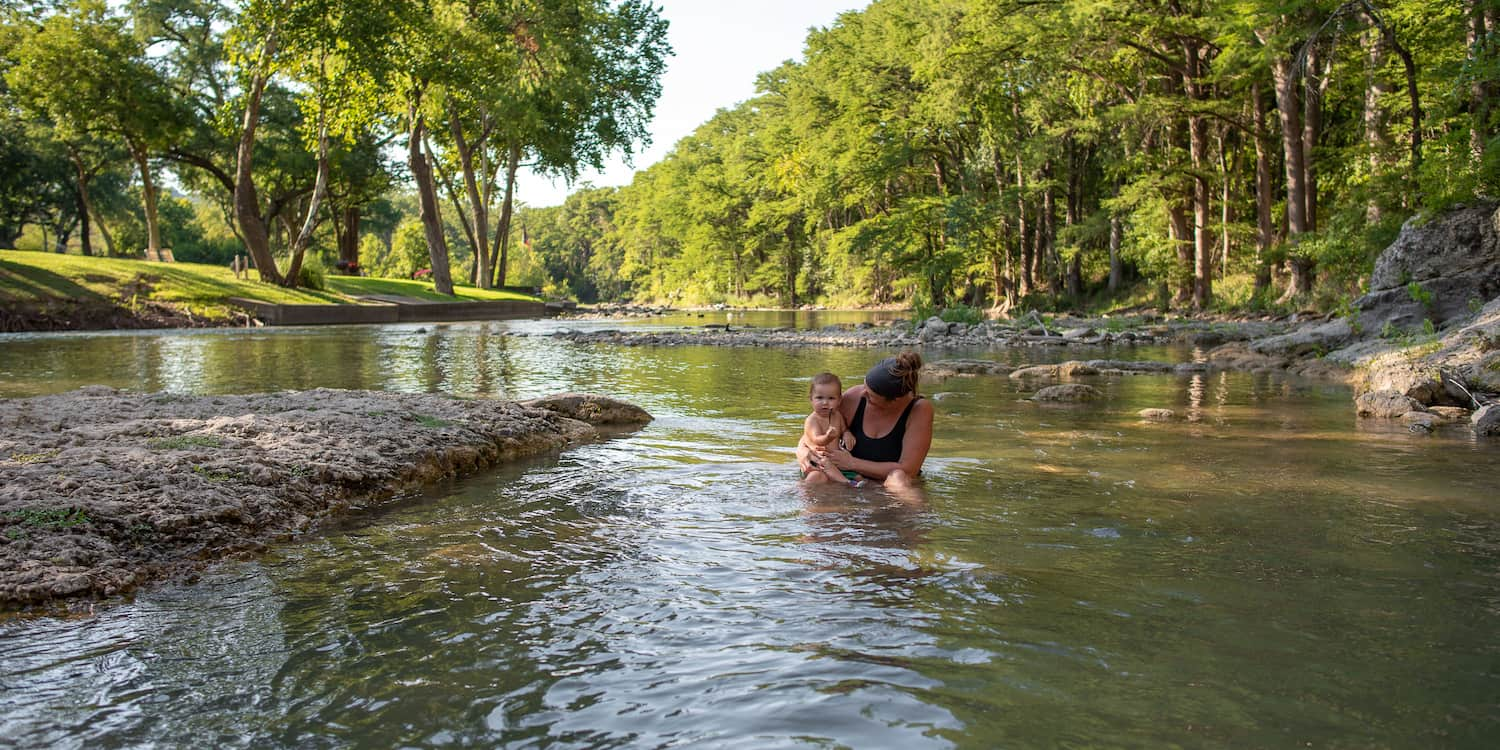 A mother and her smiling infant relaxing in the knee-deep water of the Guadalupe River at Camp Fimfo Texas Hill Country