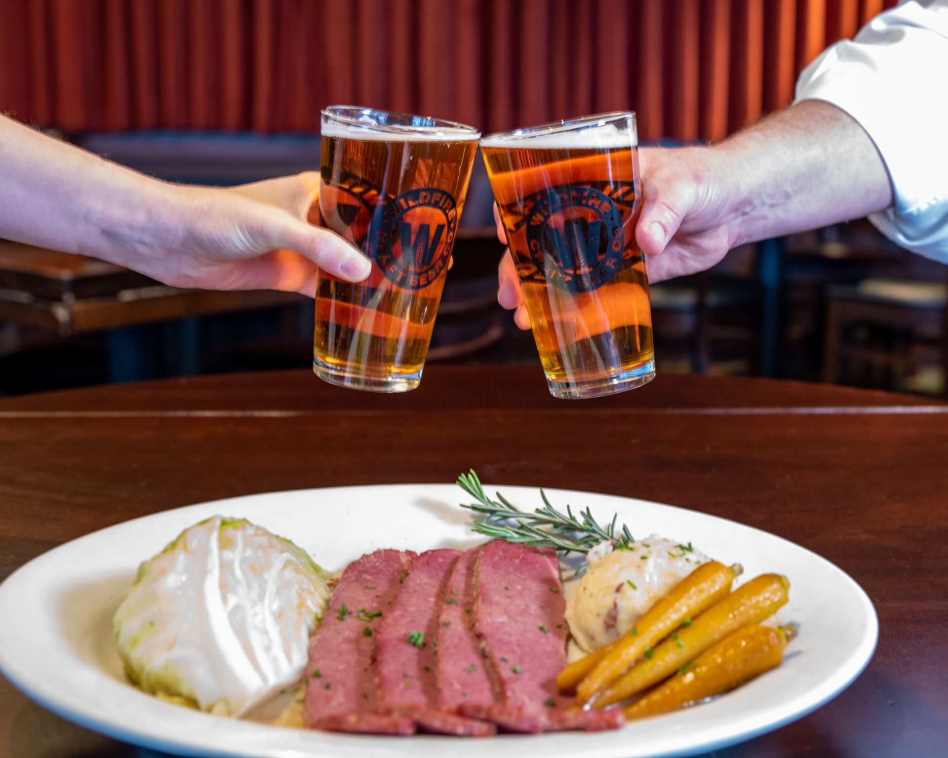 Two guests clink Wildfire Craft Beer pints over a plate of corned beef with cabbage, carrots, and mashed potatoes in a warmly lit dining room.