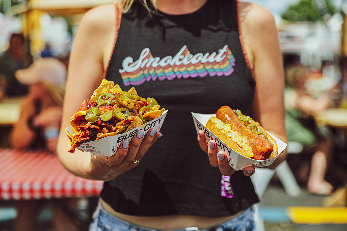 girl wearing smokeout shirt holdaing nachos and a sausage.