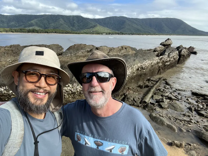 A selfie of two men in hats on the rocks, with the headland behind