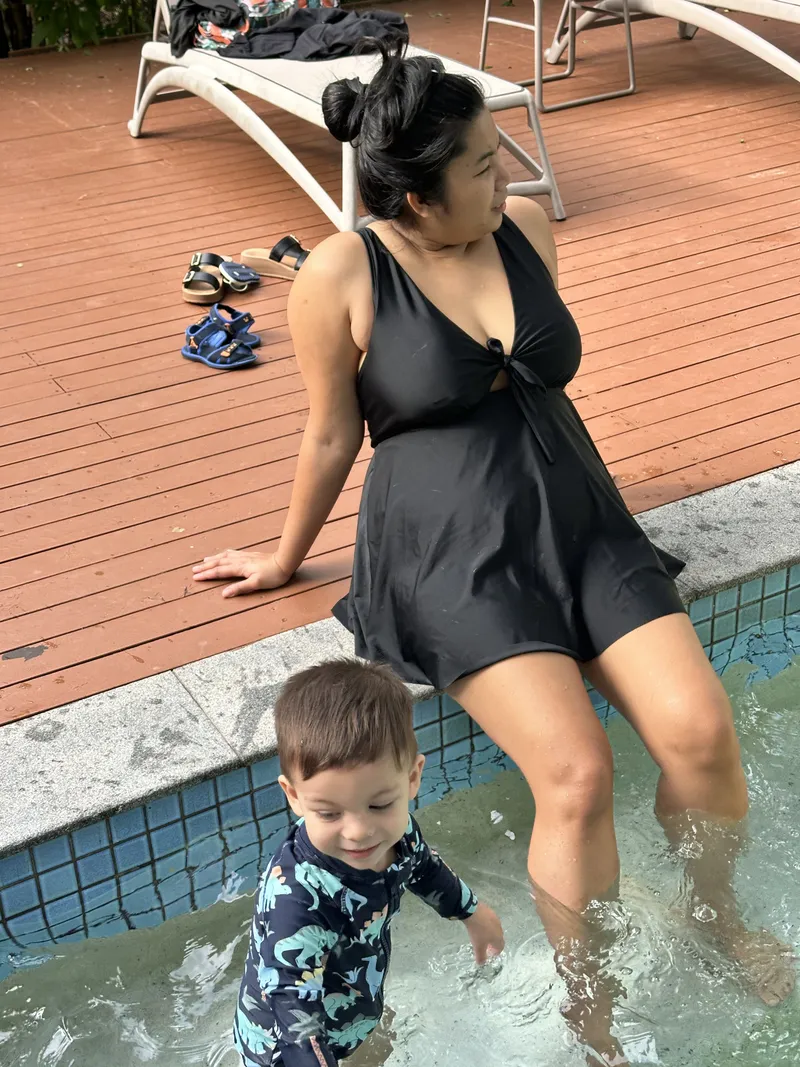 A toddler in a dinosaur swimsuit in the shallow end, with a woman sitting on the edge