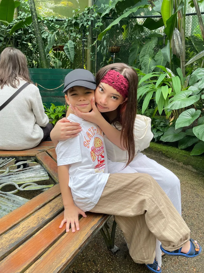 A girl in a headband hugging a boy on a bench in the glasshouse