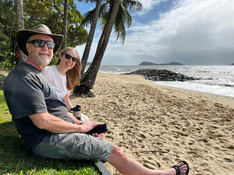 A man in a hat and a woman sitting on the grass above the beach, with palms behind