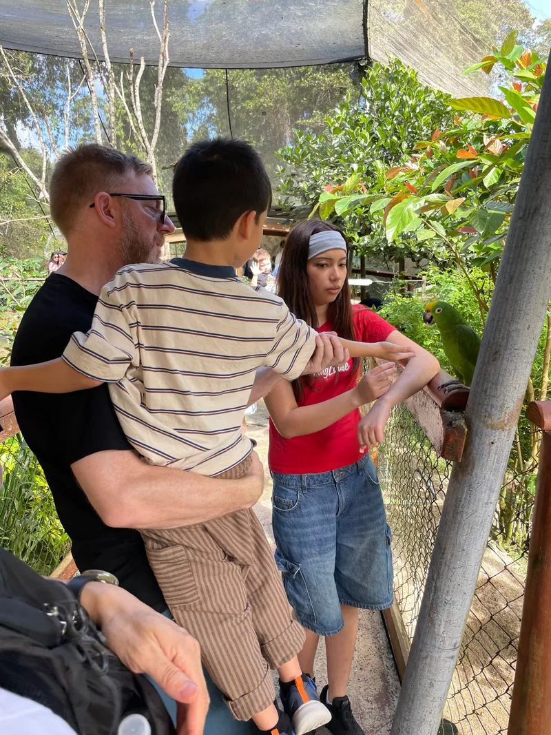 A man holding a boy up to a parrot on the railing, with the girl beside them