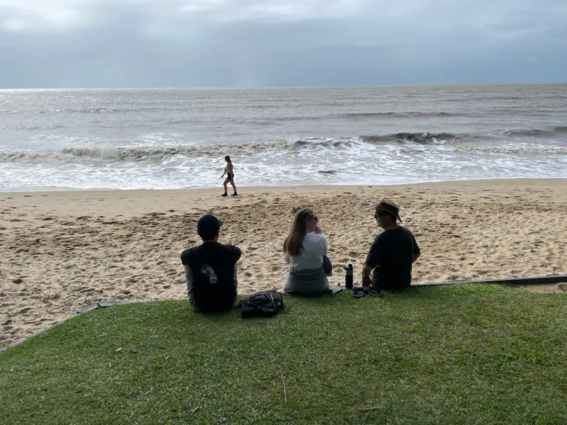 Three people sitting on the grass edge above the beach, watching the waves
