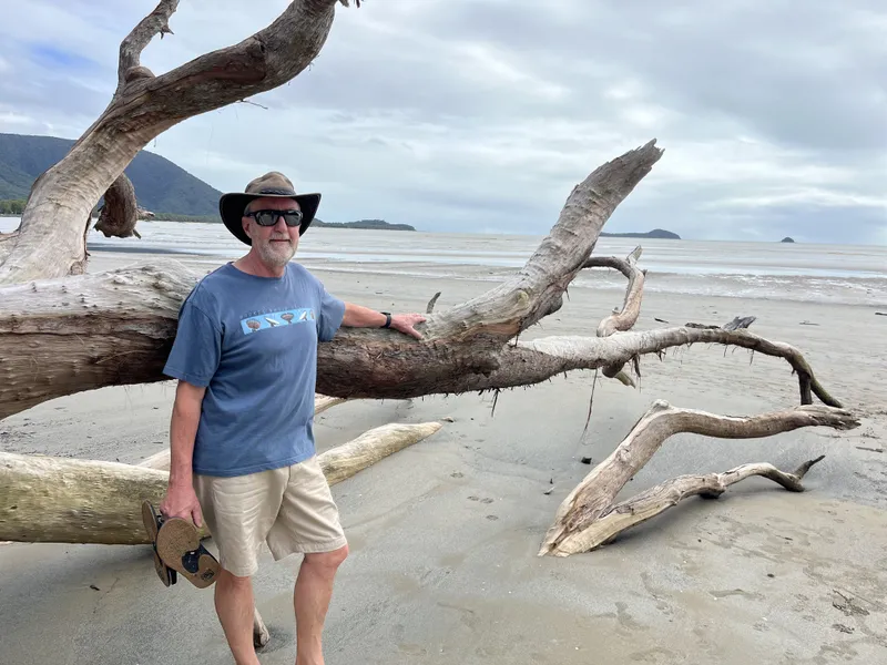 A man in a hat leaning on a huge driftwood trunk on the sand