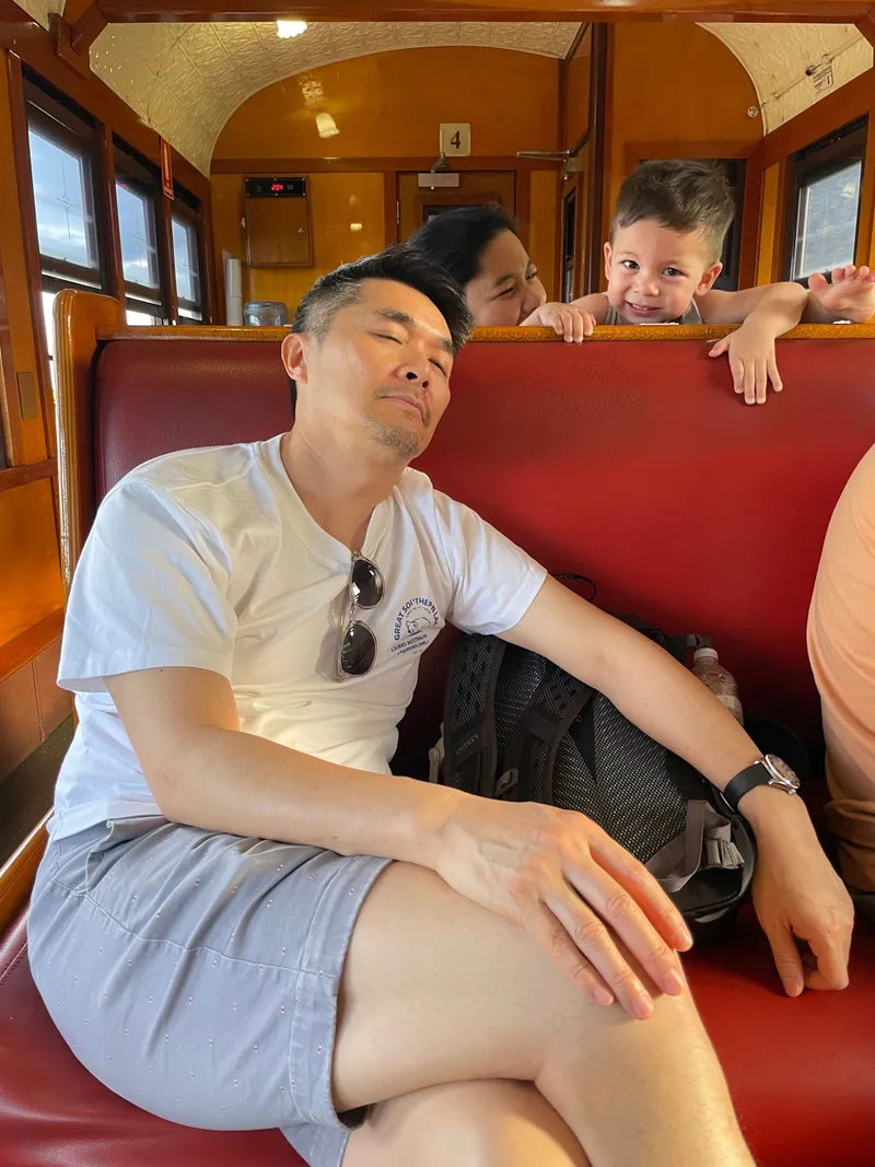 A man dozing on the train, with a boy peeking over the seat behind