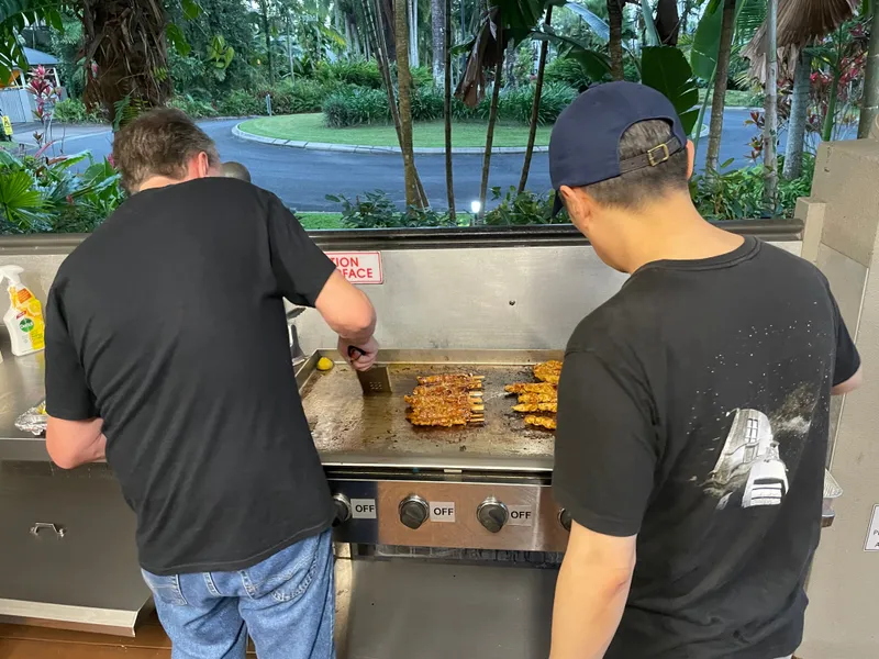Two men cooking skewers at the barbecue, with the gardens beyond