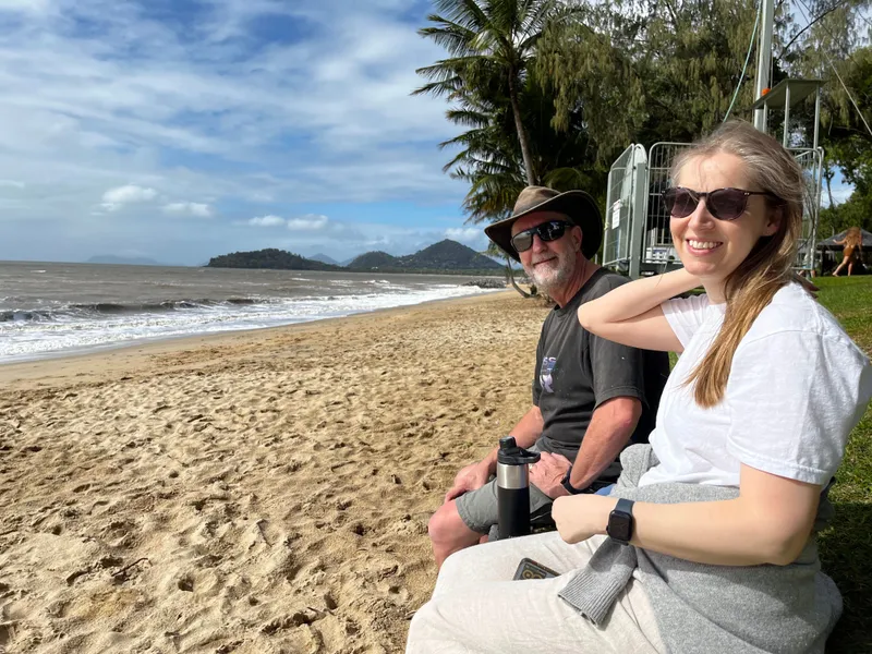 A man in a hat and a woman sitting on the grass above the beach