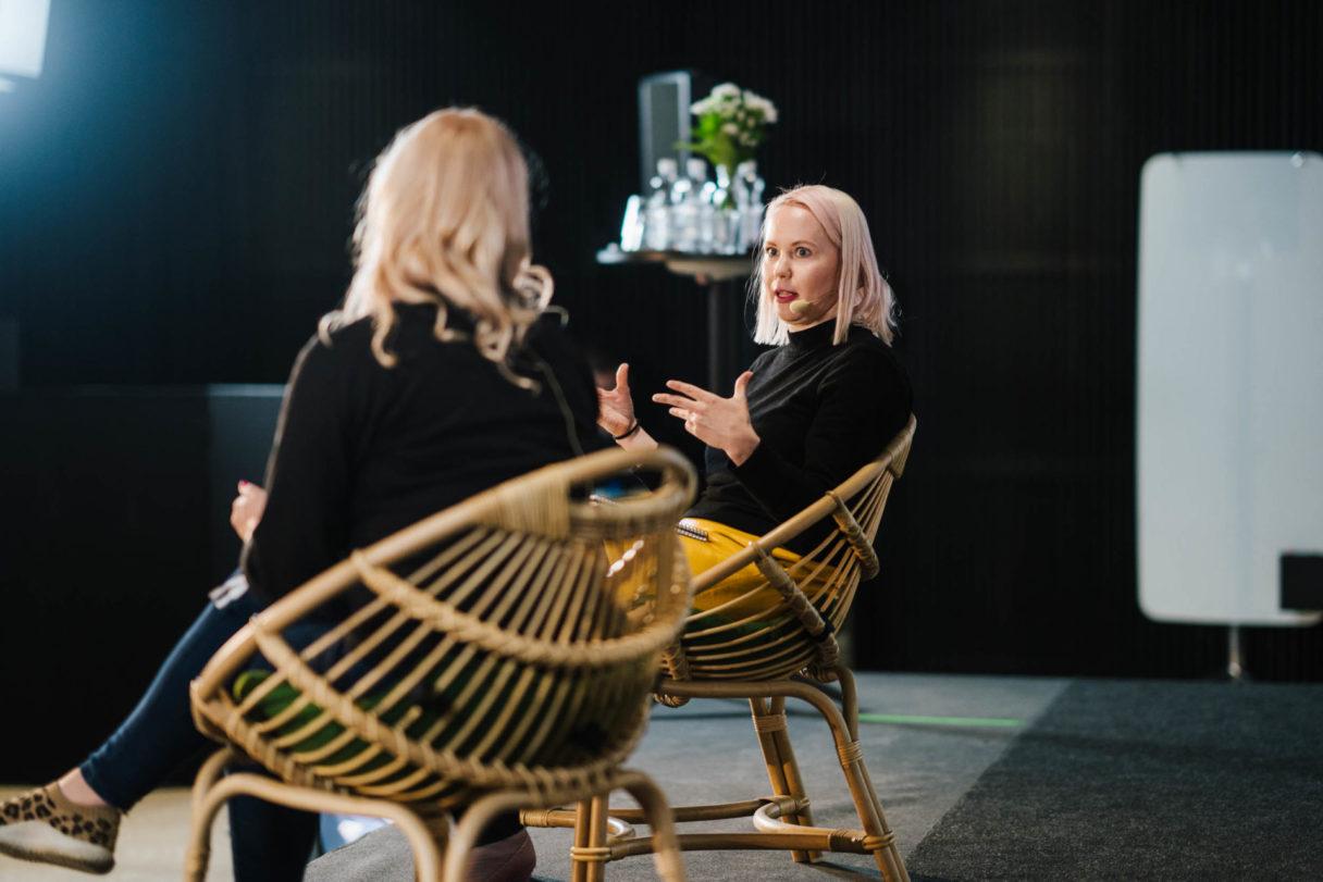 Keskustelu lavalla rottinkituoleissa, taustalla tarjoilupöytä ja tapahtumavalaistus.