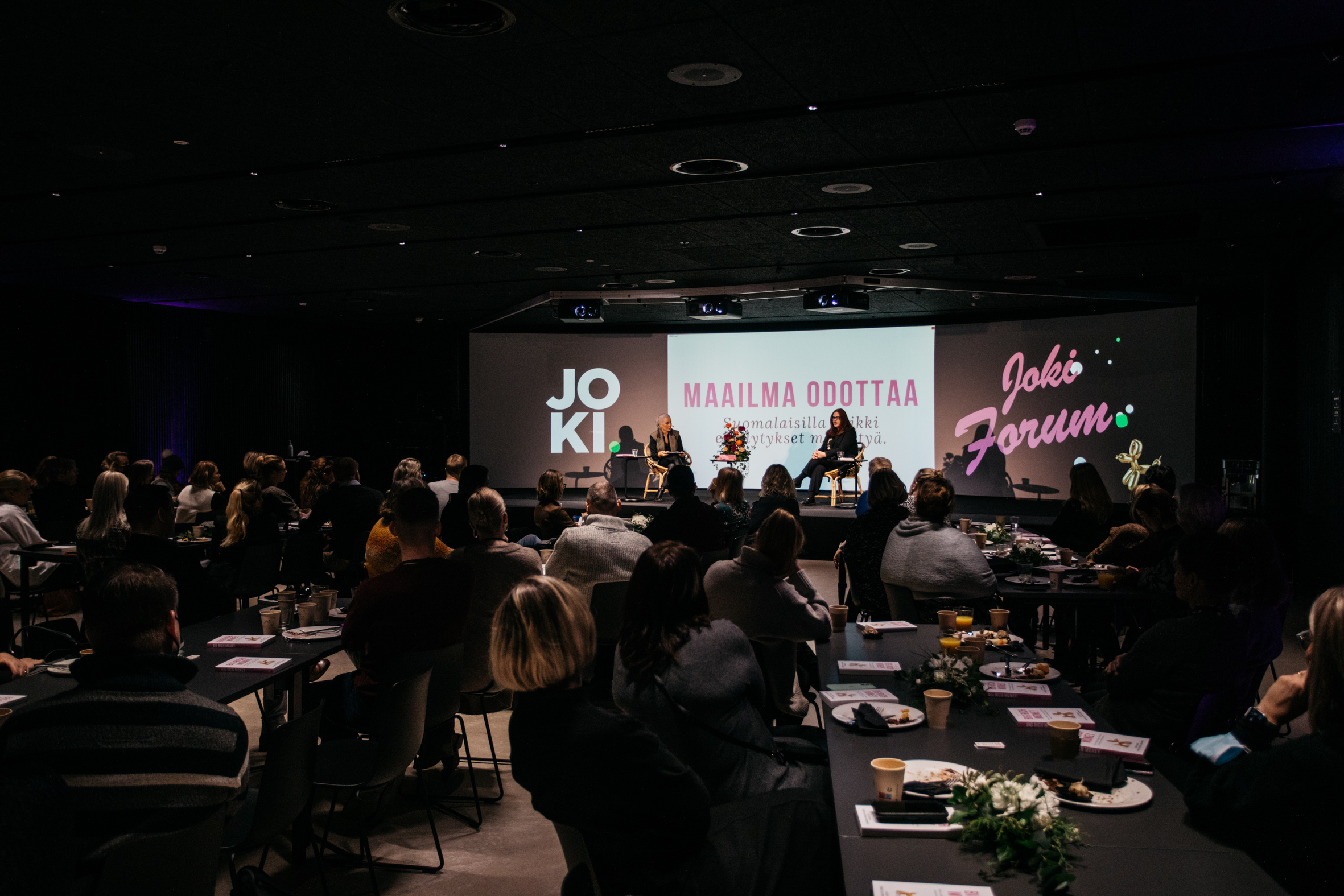 Tapahtuma Vierailukeskus Joen Cave-teatterissa. Kaarevalla näytöllä teksti Maailma odottaa, lavalla kaksi ihmistä keskustelemassa.